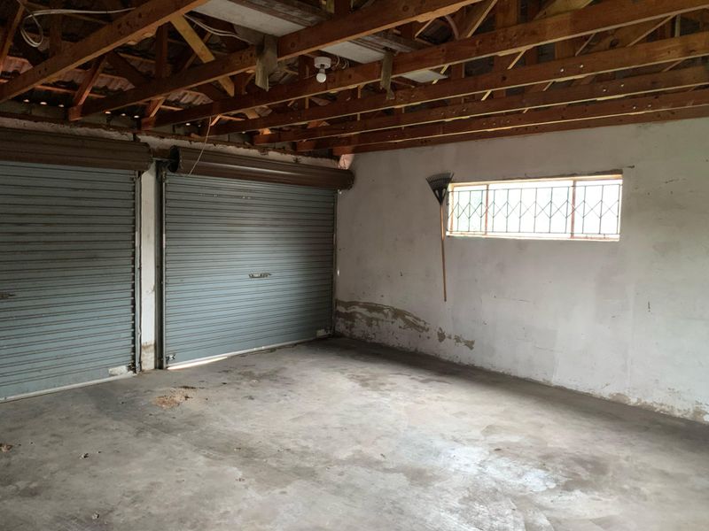A spacious double garage featuring exposed timber roof trusses, concrete flooring, and dual roller shutter doors, requiring maintenance and cosmetic updates.