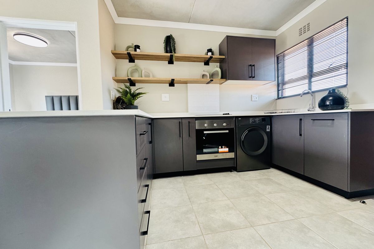 Modern kitchen featuring dark cabinetry, built-in oven, integrated washing machine, open shelving, and light-toned tiled flooring.