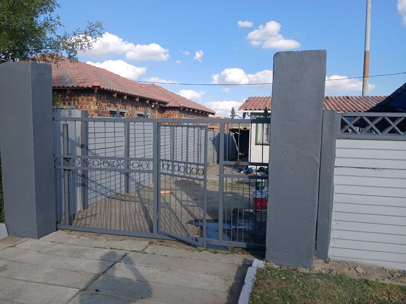A secure front entrance featuring a grey metal gate, concrete pillars, and perimeter walling, providing access to the property's driveway and yard area.