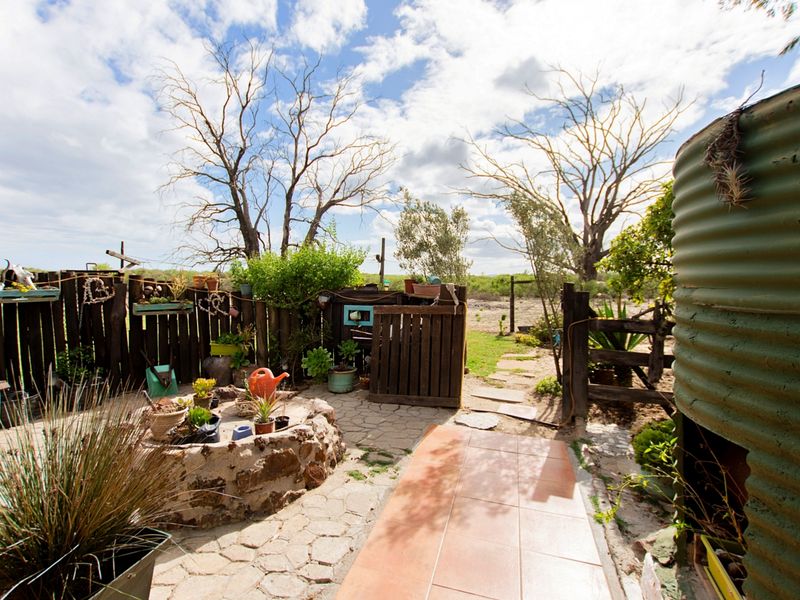 A rustic outdoor garden area featuring a stone-clad water feature, corrugated metal water tank, and paved pathways enclosed by timber fencing.