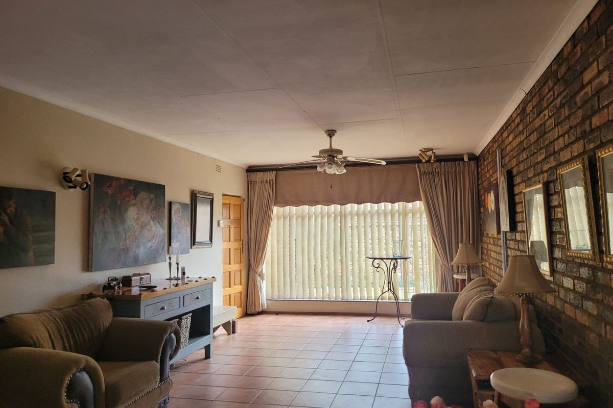 A spacious living room featuring an exposed brick feature wall, tiled flooring, and a ceiling fan, with large windows providing natural light.