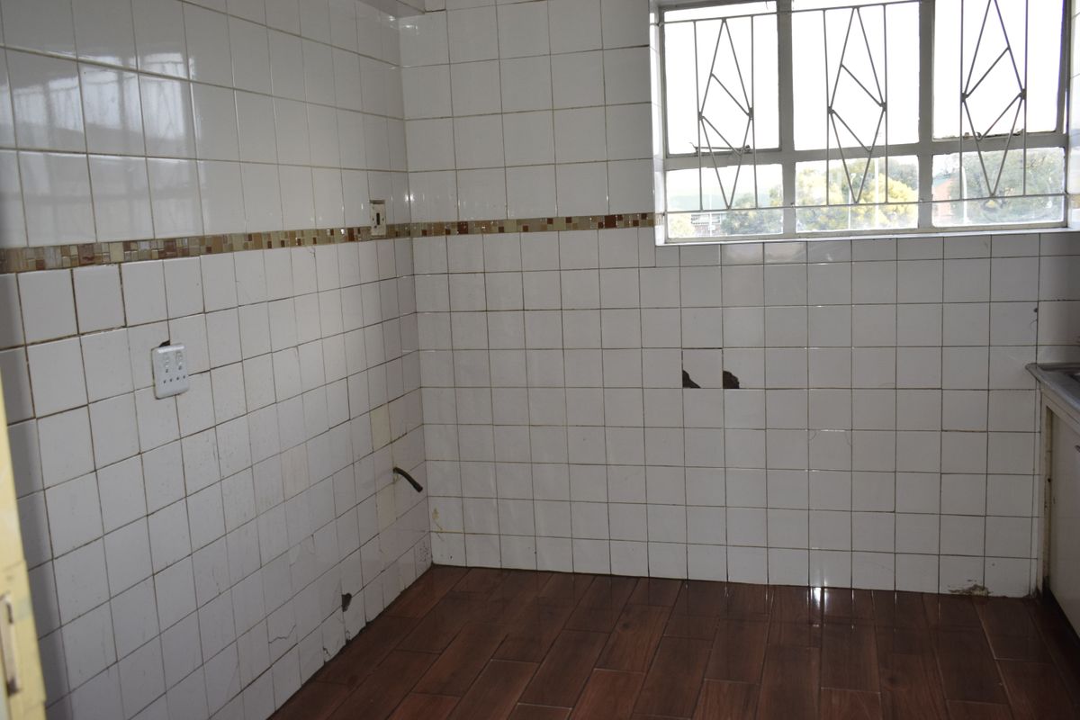 An empty kitchen area featuring tiled walls with some damage, wood-look floor tiles, and a window with security bars.