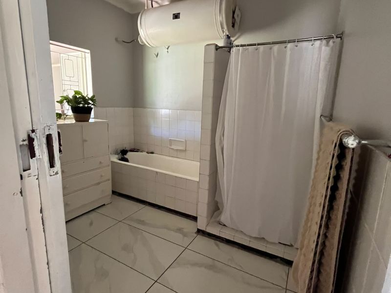 A functional bathroom featuring a built-in bathtub, separate shower enclosure, and wall-mounted geyser, finished with tiled flooring and wall splashbacks.
