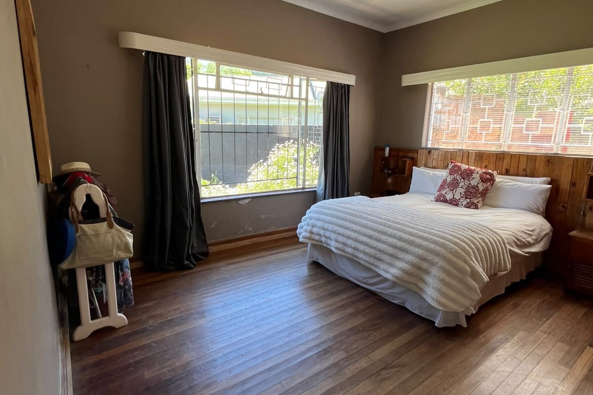 Spacious bedroom featuring warm timber flooring, a built-in wooden headboard, and large windows with security bars, offering a classic foundation for updates.