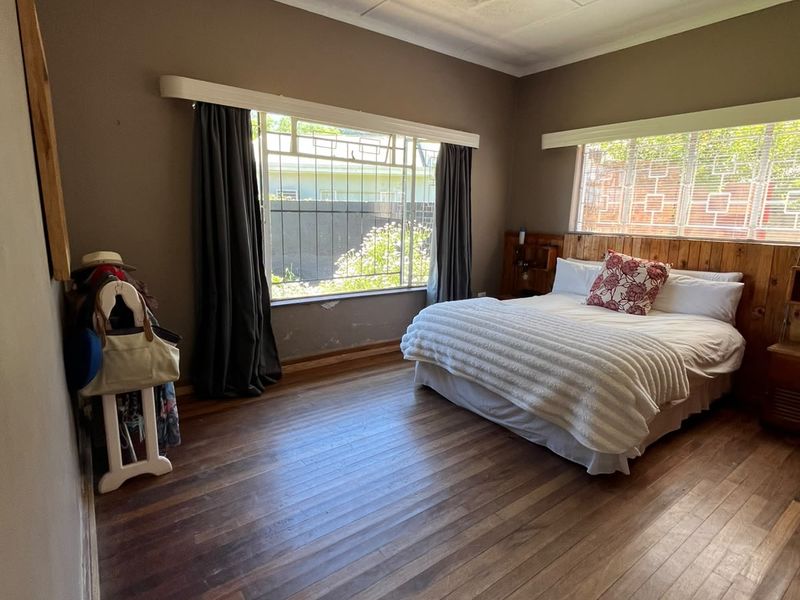 Spacious bedroom featuring warm timber flooring, a built-in wooden headboard, and large windows with security bars, offering a classic foundation for updates.