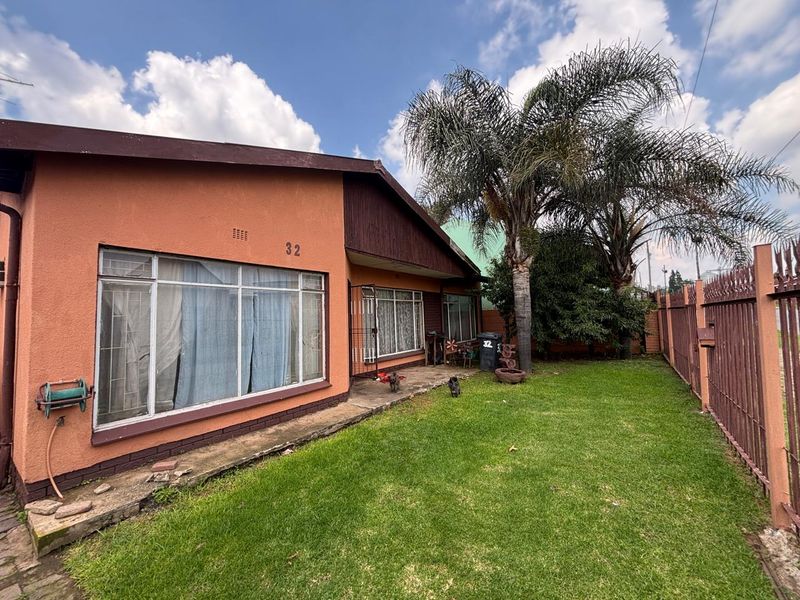 Front exterior view of a single-story house featuring a lawn, metal security fencing, and large windows with a stucco finish.