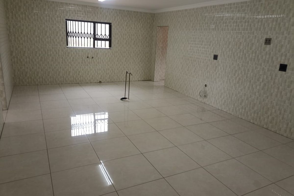 An empty, tiled kitchen space featuring wall-to-wall tiling, plumbing connections, and a window with a security gate.