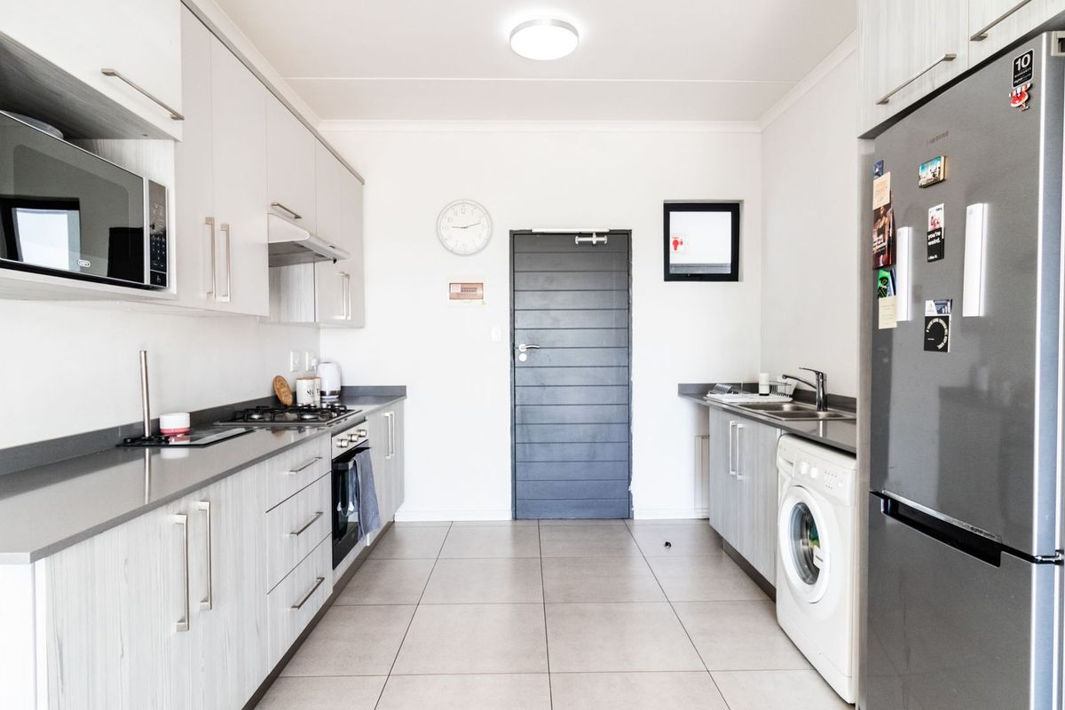 Modern galley-style kitchen featuring light wood-grain cabinetry, stone-look countertops, integrated appliances, and tiled flooring with dedicated laundry plumbing.