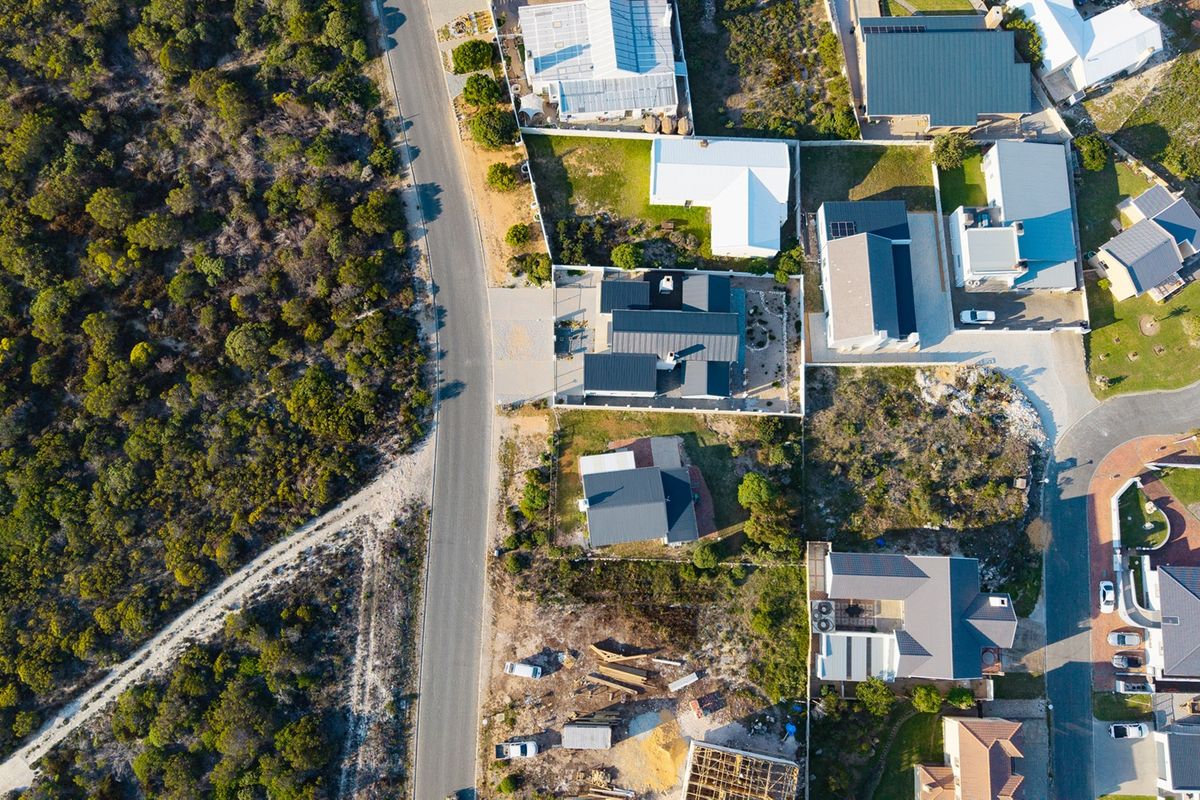 An aerial view of a residential neighborhood showing multiple properties, metal roof structures, and surrounding natural vegetation and road infrastructure.