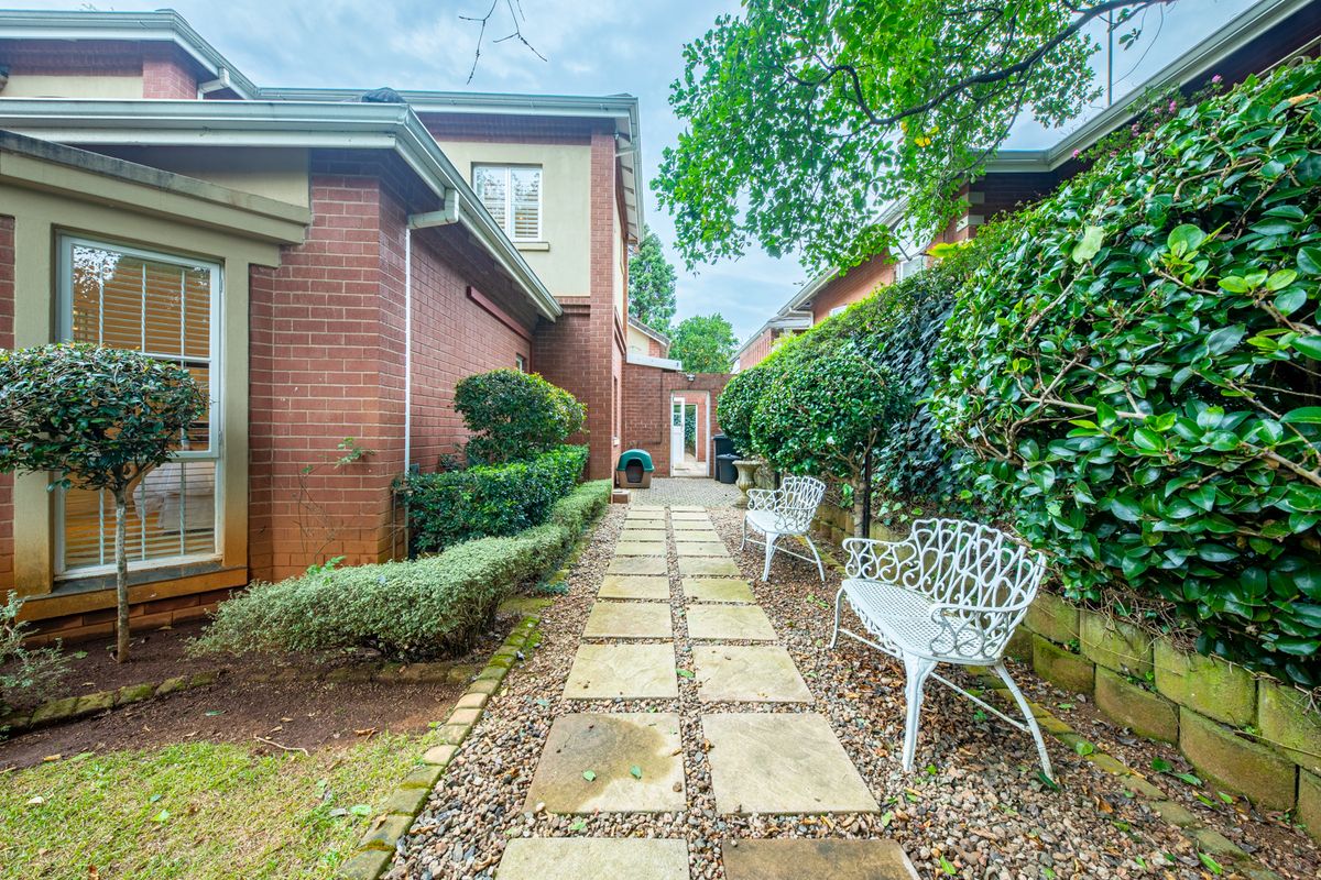A well-maintained rear garden path featuring stone pavers, brick exterior walls, and established green hedging along a retaining wall.