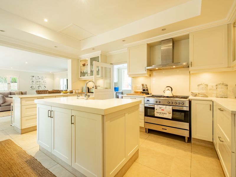 A bright open-plan kitchen featuring a central island, stone countertops, stainless steel gas stove, and ample built-in cabinetry, seamlessly flowing into the living area.