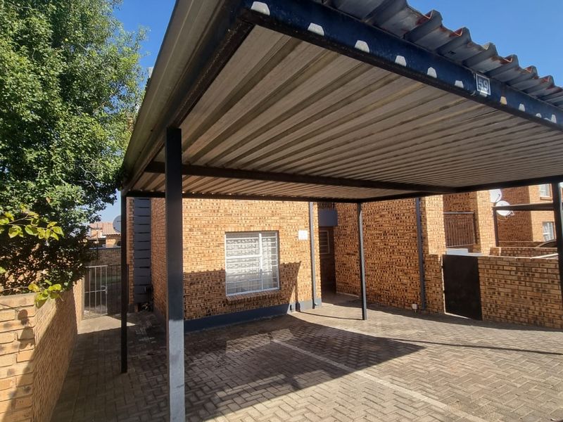 A covered carport with a corrugated metal roof and brick paving, situated in front of a facebrick apartment unit.
