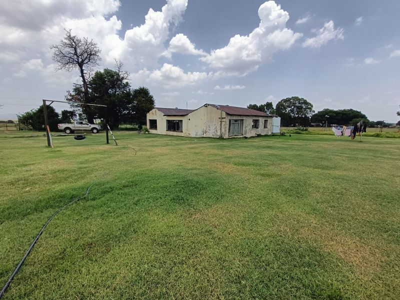 A view of the rear exterior of a farm dwelling set on a large grassy plot with visible signs of exterior wear.