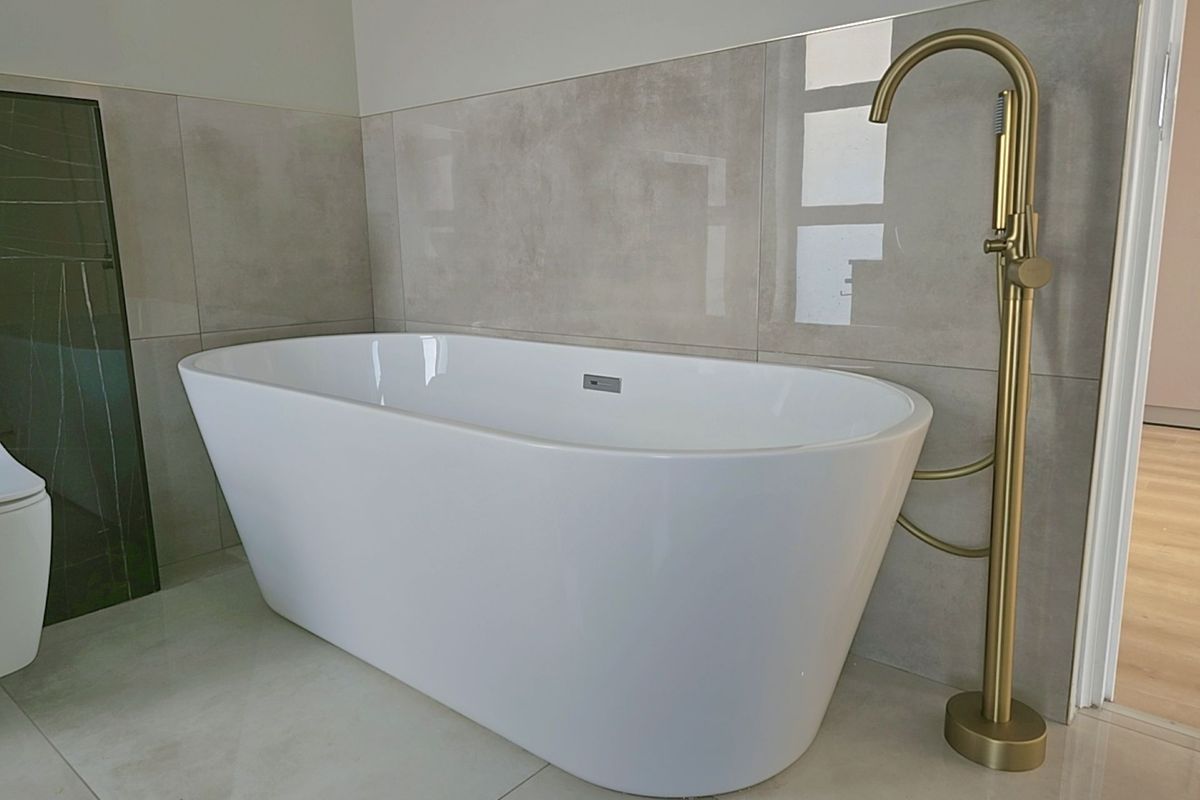 Modern bathroom featuring a sleek freestanding bathtub with a contemporary floor-mounted brass faucet and neutral large-format wall and floor tiling.