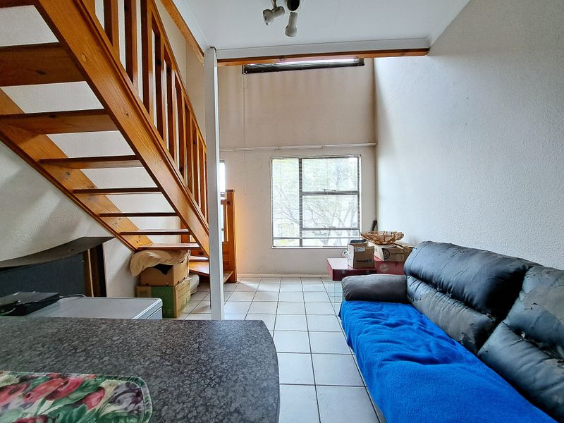 A living area featuring a wooden staircase, tiled flooring, and a large window, offering a double-volume space with potential for modernization.