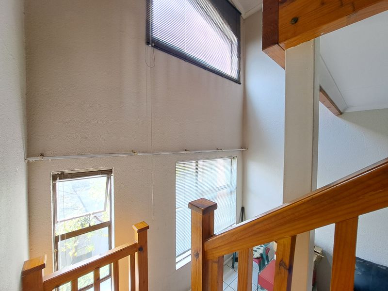 A double-volume hallway area featuring a wooden staircase railing, high ceilings, and multiple windows with fitted venetian blinds.