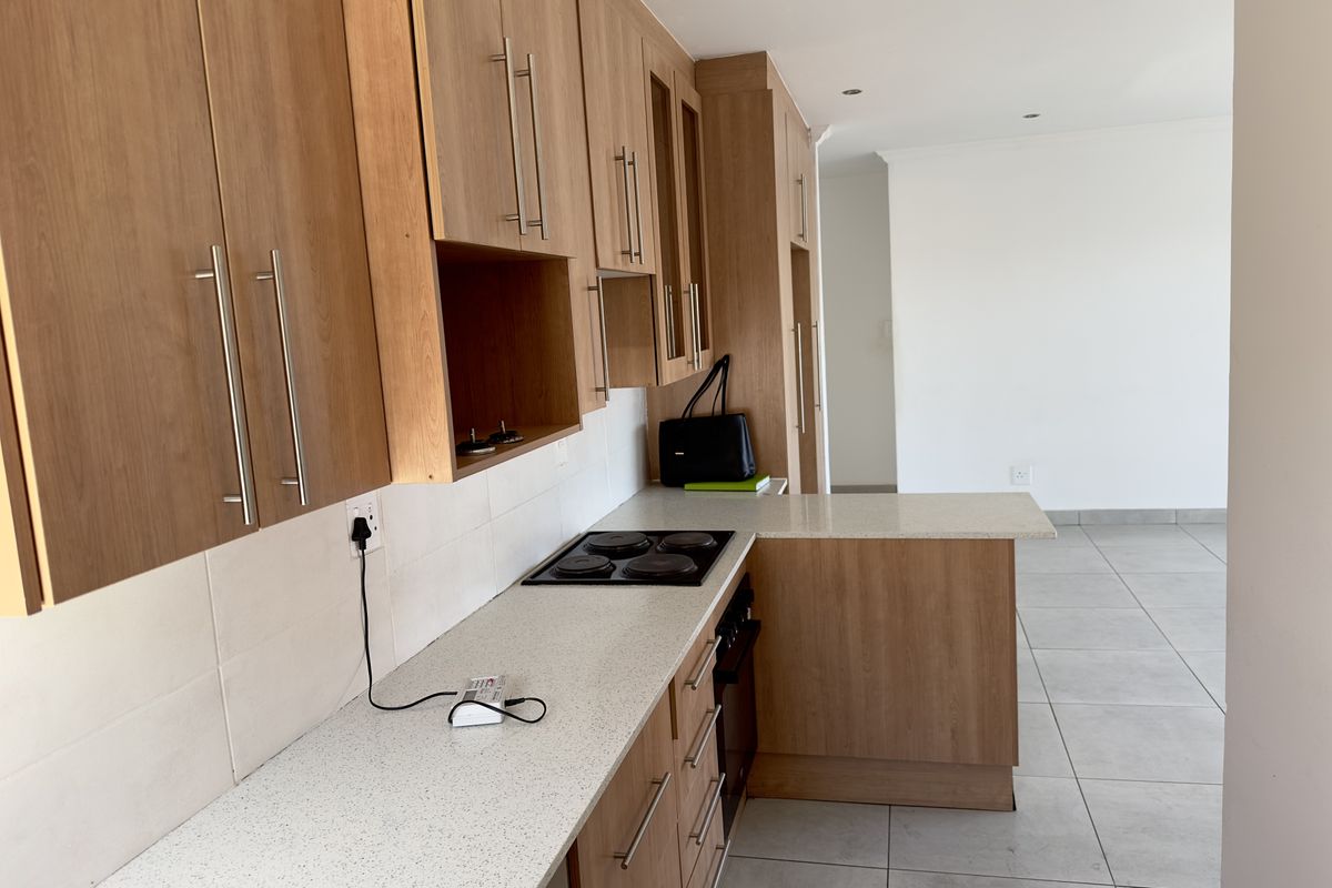 Modern kitchen featuring light wood cabinetry, stone-look countertops, a built-in electric hob, and an integrated oven, flowing into an open-plan living area.