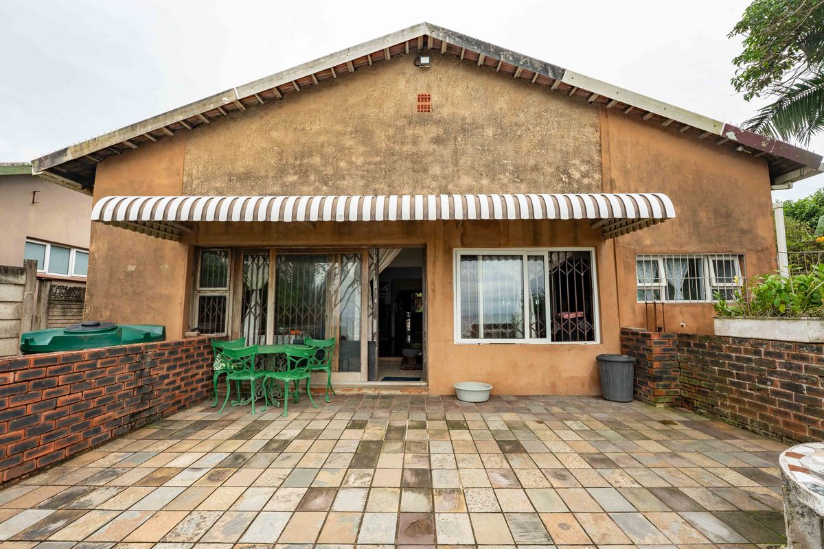 Rear exterior view featuring a paved patio area, sliding glass doors, security gates, and a weathered facade with a striped awning.
