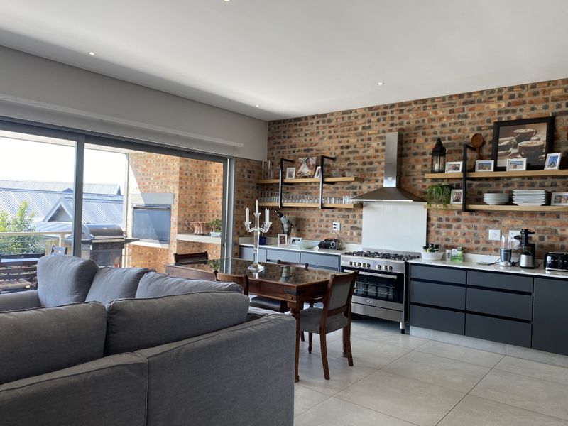 Modern open-plan living and kitchen area featuring an exposed brick feature wall, sleek cabinetry, and seamless flow to a covered balcony with built-in braai.