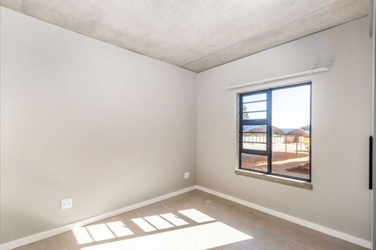 A bright, empty bedroom featuring a modern exposed concrete ceiling, neutral walls, and a large window with metal frames.