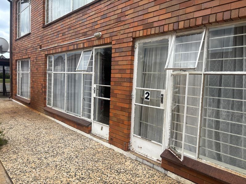 Exterior view of an apartment building featuring face-brick facade, paved walkway, and multiple glass-paneled entrance doors with security bars.