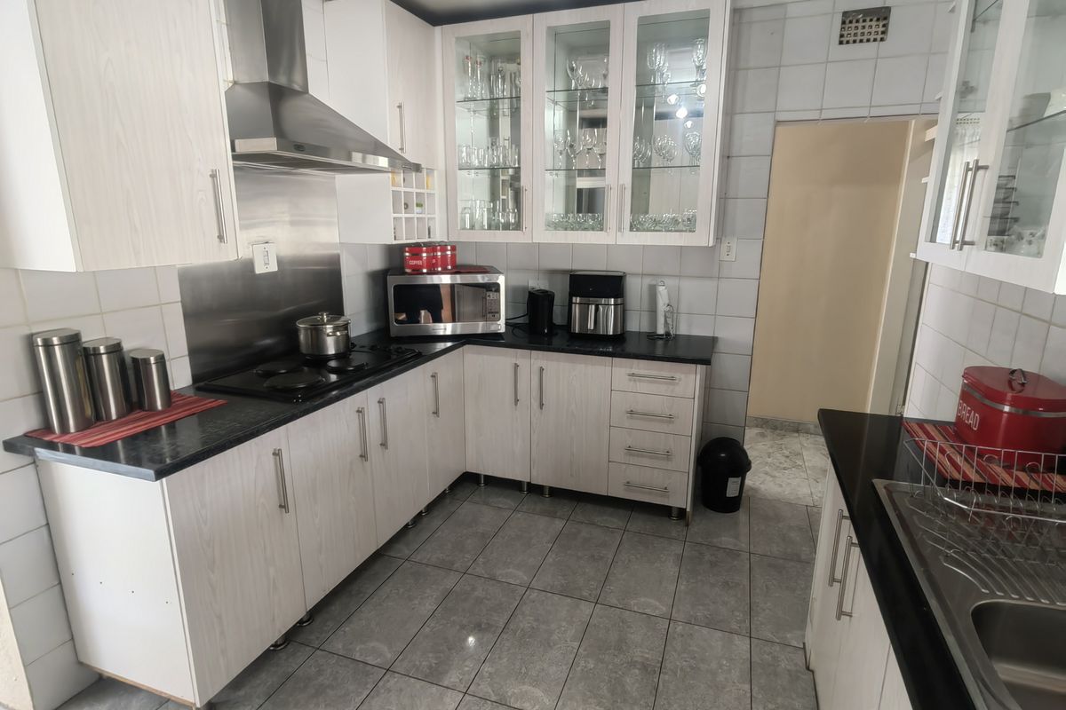 A functional kitchen featuring light wood-grain cabinetry, black countertops, a stainless steel extractor fan, and tiled flooring with ample storage space.
