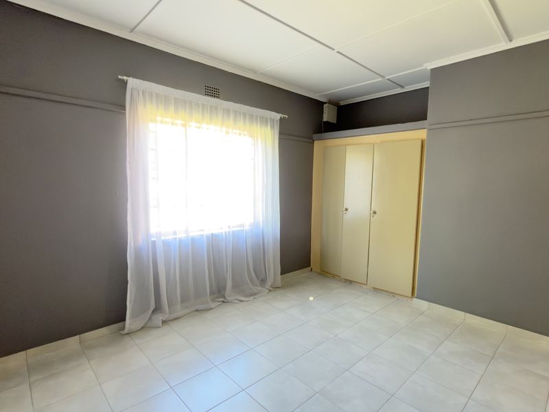 A bright, empty bedroom featuring tiled flooring, built-in cupboards, and a window with security bars.