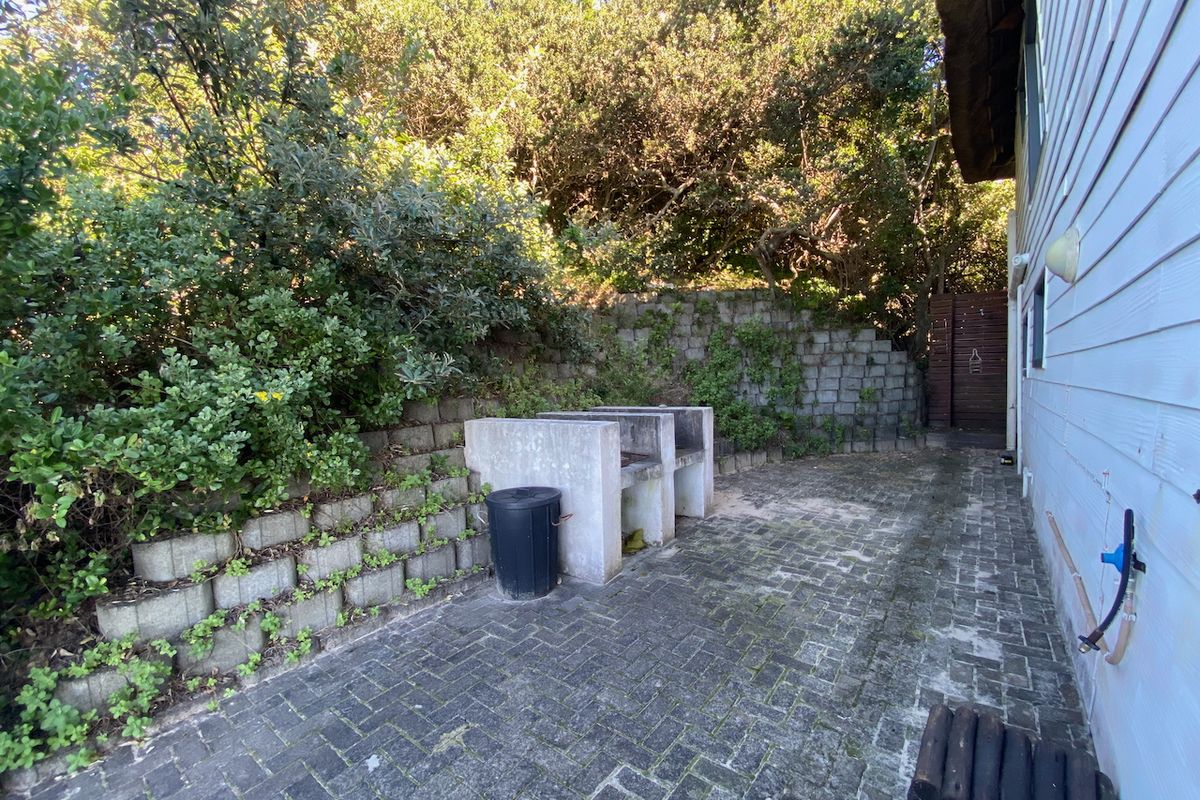 Rear exterior area featuring a paved patio, built-in braai, and a tiered retaining wall set against a backdrop of mature trees.