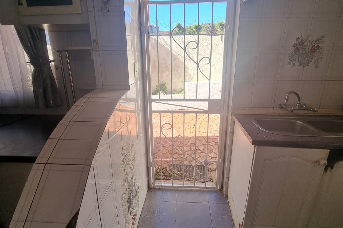 A dated kitchen area featuring tiled surfaces, built-in cabinetry, and a double bowl sink, with direct access to the exterior via a security gate.