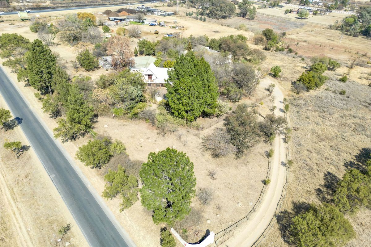 Aerial view of a small holding property featuring a gravel driveway, established trees, and perimeter fencing with direct access to the main road.