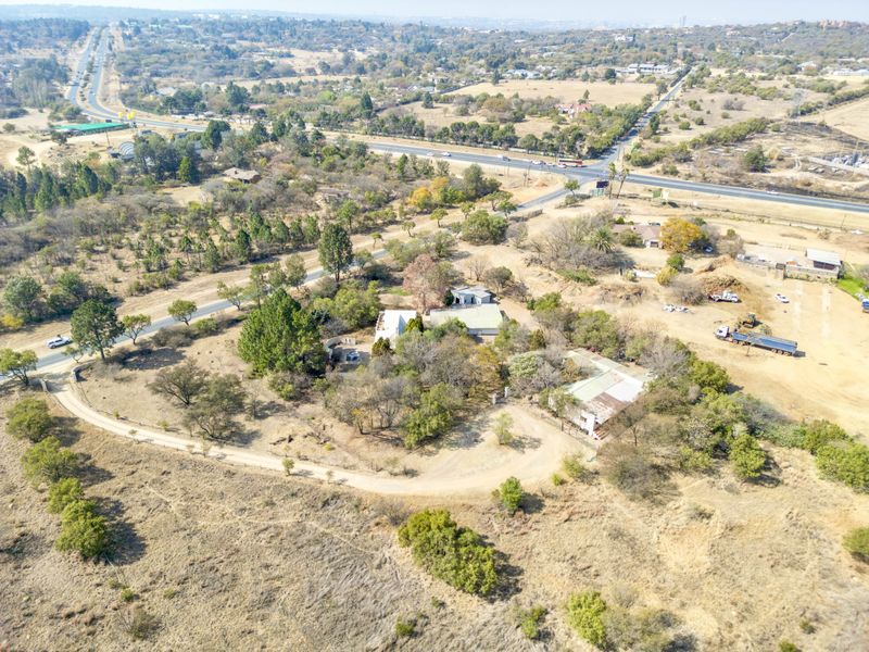 Aerial view of a small holding property featuring multiple structures, mature trees, and a gravel driveway with surrounding landscape.
