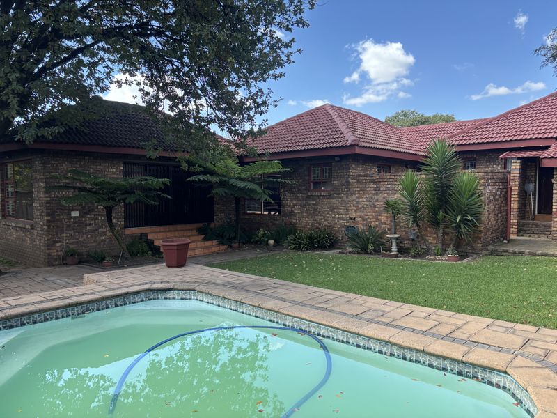 A well-maintained outdoor pool area featuring a paved deck, established garden, and a classic face brick house exterior with a tiled roof.
