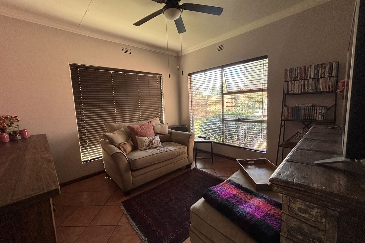 A cozy living room featuring terracotta floor tiles, a ceiling fan, and large windows with venetian blinds, offering a comfortable and functional living space.