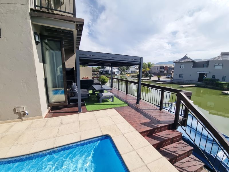 Waterfront patio featuring timber decking, a modern metal pergola, built-in braai, and direct access to a private swimming pool overlooking the canal.