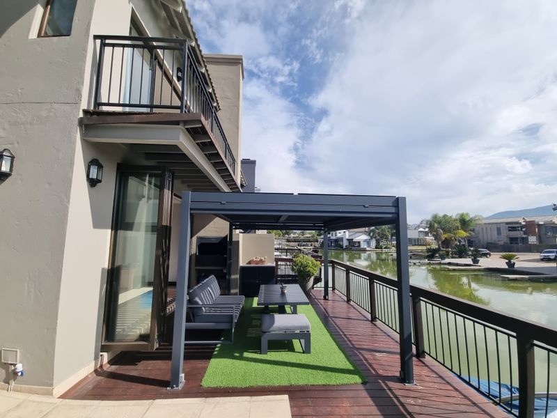 Waterfront patio featuring timber decking, a modern metal pergola, built-in braai, and bi-fold doors providing seamless indoor-outdoor flow.