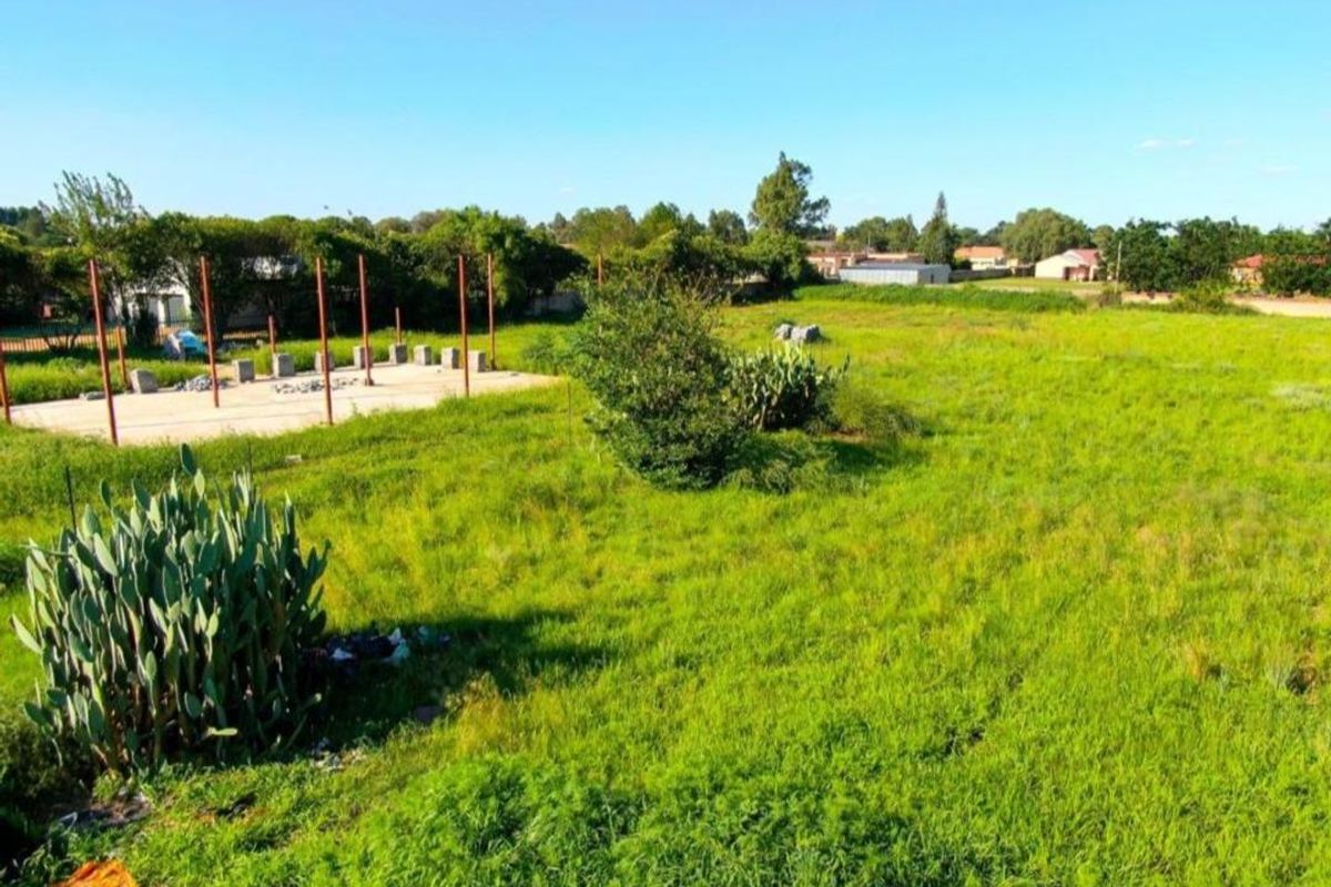 A vacant residential plot featuring an unfinished concrete foundation with metal support poles, surrounded by open grassy land and established vegetation.