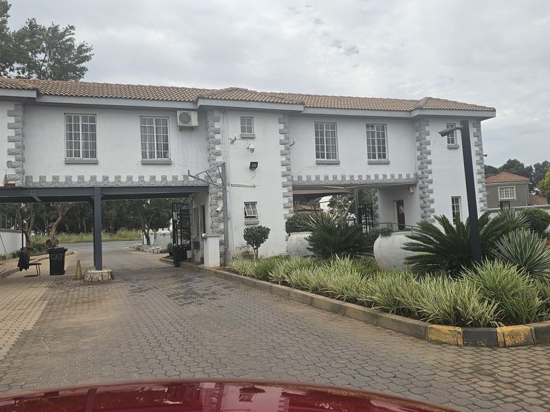 View of the apartment complex entrance gatehouse featuring a paved driveway, security access point, and established landscaped garden beds.