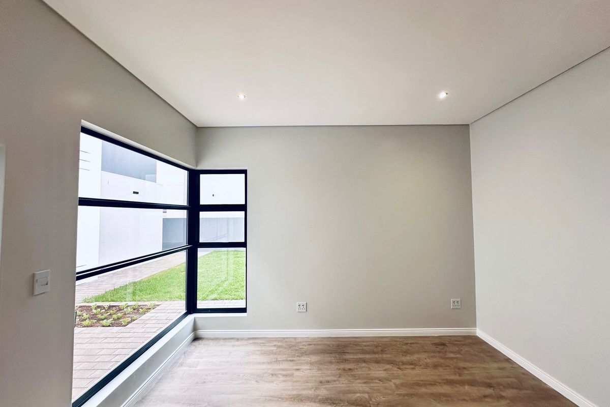 A bright, modern, and empty bedroom featuring neutral walls, laminate flooring, and large black-framed windows overlooking the exterior garden area.