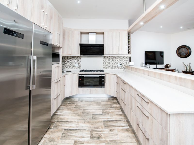Modern kitchen featuring light wood-grain cabinetry, stone countertops, mosaic backsplash, and integrated appliances, seamlessly flowing into the adjacent living area.