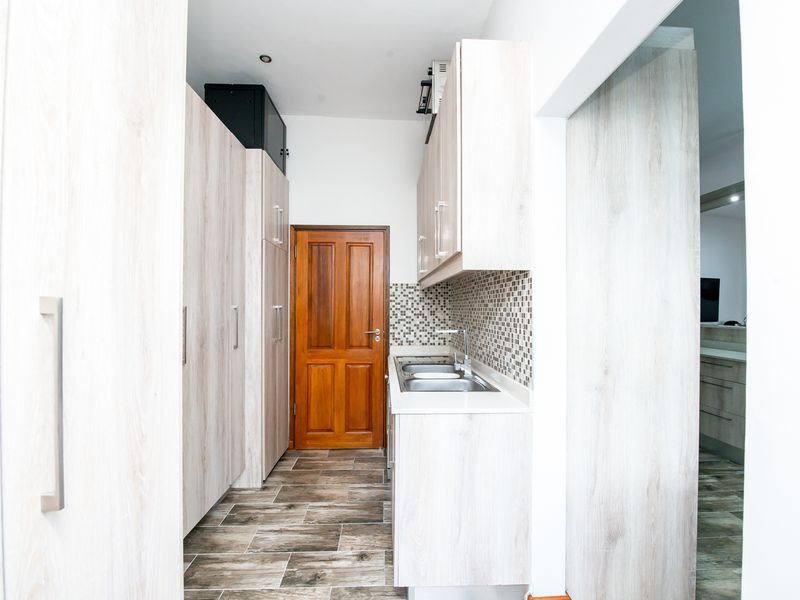 A clean and functional scullery featuring light wood-look cabinetry, a stainless steel sink, mosaic tile backsplash, and durable tiled flooring.