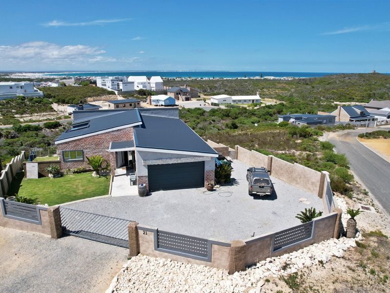 Aerial view of a modern brick house with a metal roof, gravel driveway, and secure perimeter wall in a coastal residential estate.