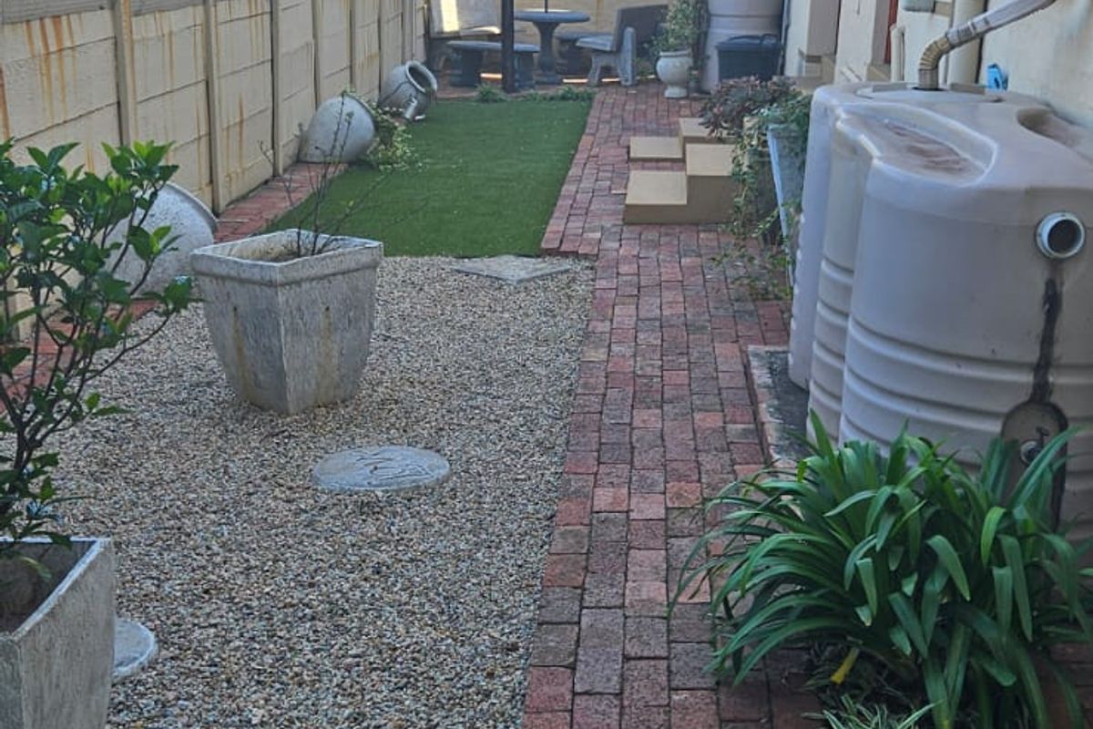A side garden area featuring brick pathways, gravel landscaping, artificial turf, and installed water storage tanks along the exterior wall.