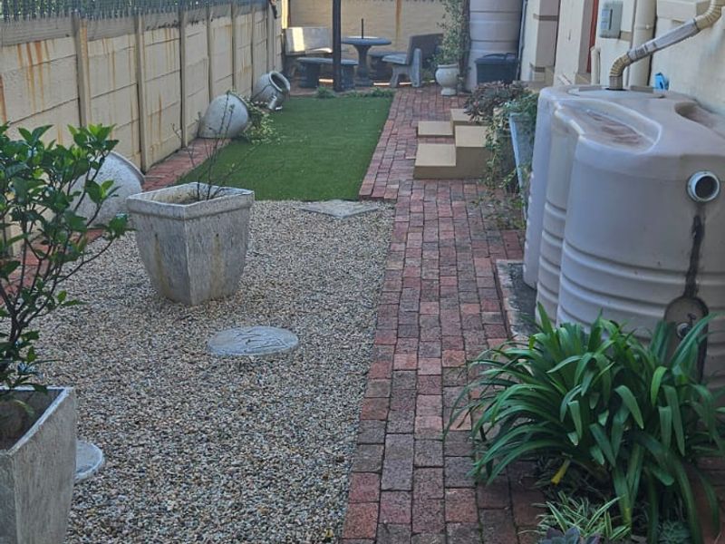 A side garden area featuring brick pathways, gravel landscaping, artificial turf, and installed water storage tanks along the exterior wall.