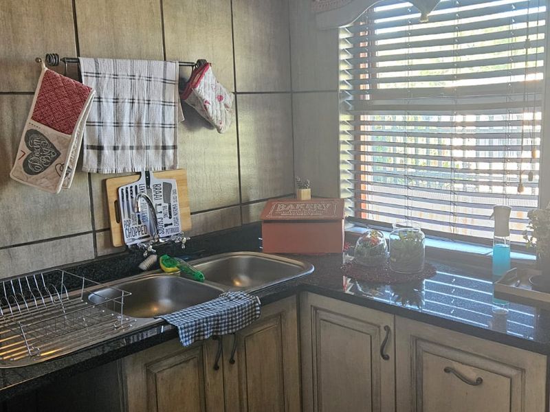 A functional scullery area featuring granite countertops, double bowl stainless steel sink, timber cabinetry, and tiled splashback with natural light from a window.