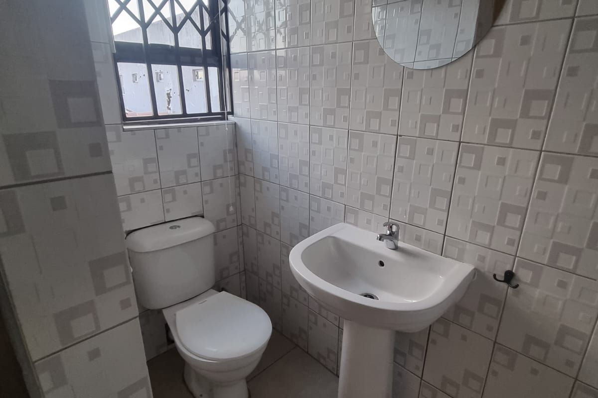 A functional bathroom featuring a pedestal basin, toilet, and fully tiled walls and floors, complete with a security-fitted window.