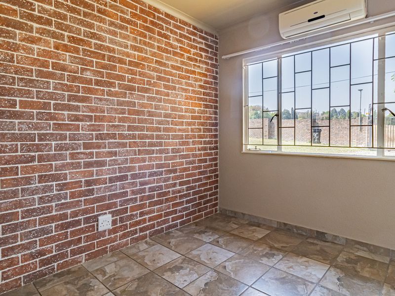 A bright, empty bedroom featuring a rustic exposed brick wall, tiled flooring, and a wall-mounted air conditioning unit with security bars on the window.