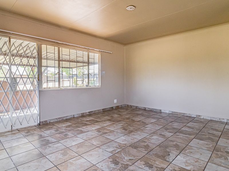 A spacious, empty living area featuring neutral tiled flooring, a sliding door with security gate, and large windows with burglar bars.