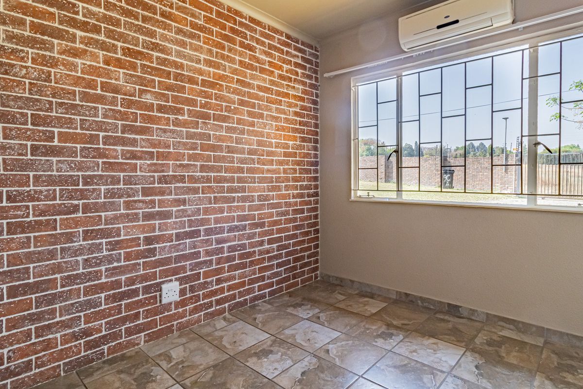 A bright, empty bedroom featuring a rustic exposed brick wall, tiled flooring, and a wall-mounted air conditioning unit with security bars on the window.