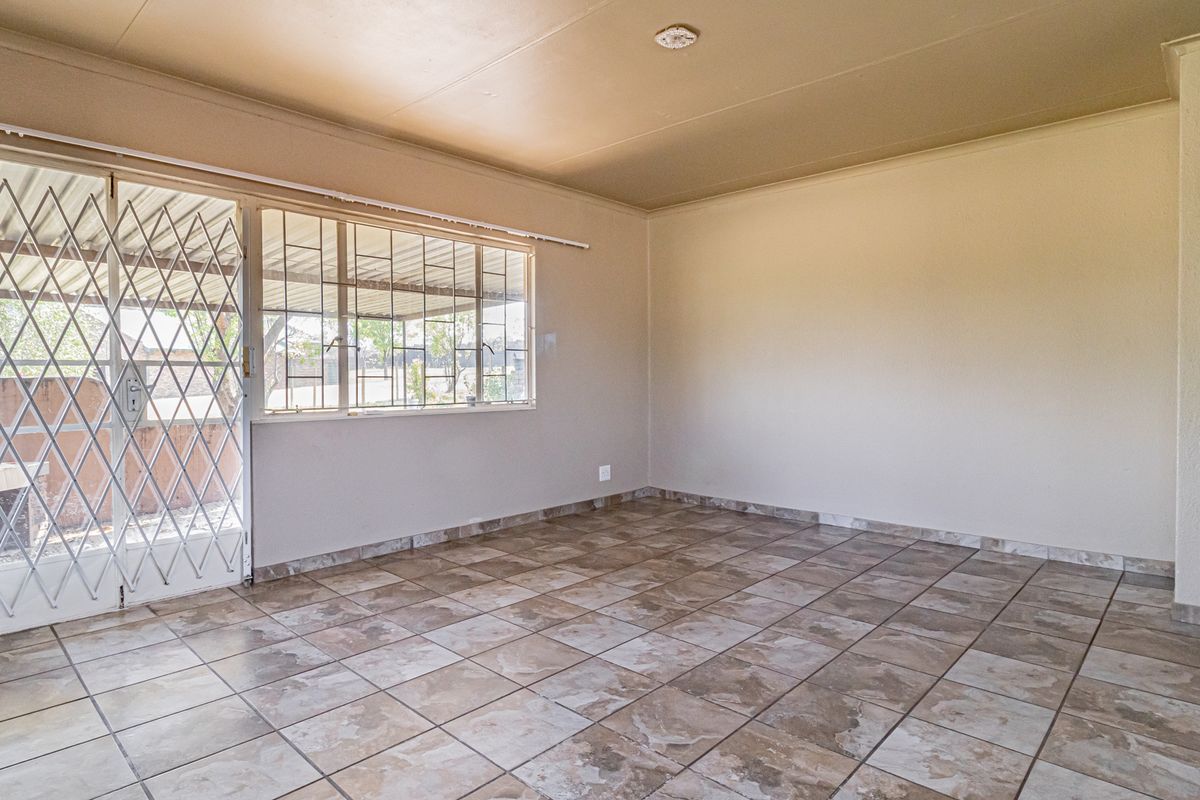 A spacious, empty living area featuring neutral tiled flooring, a sliding door with security gate, and large windows with burglar bars.