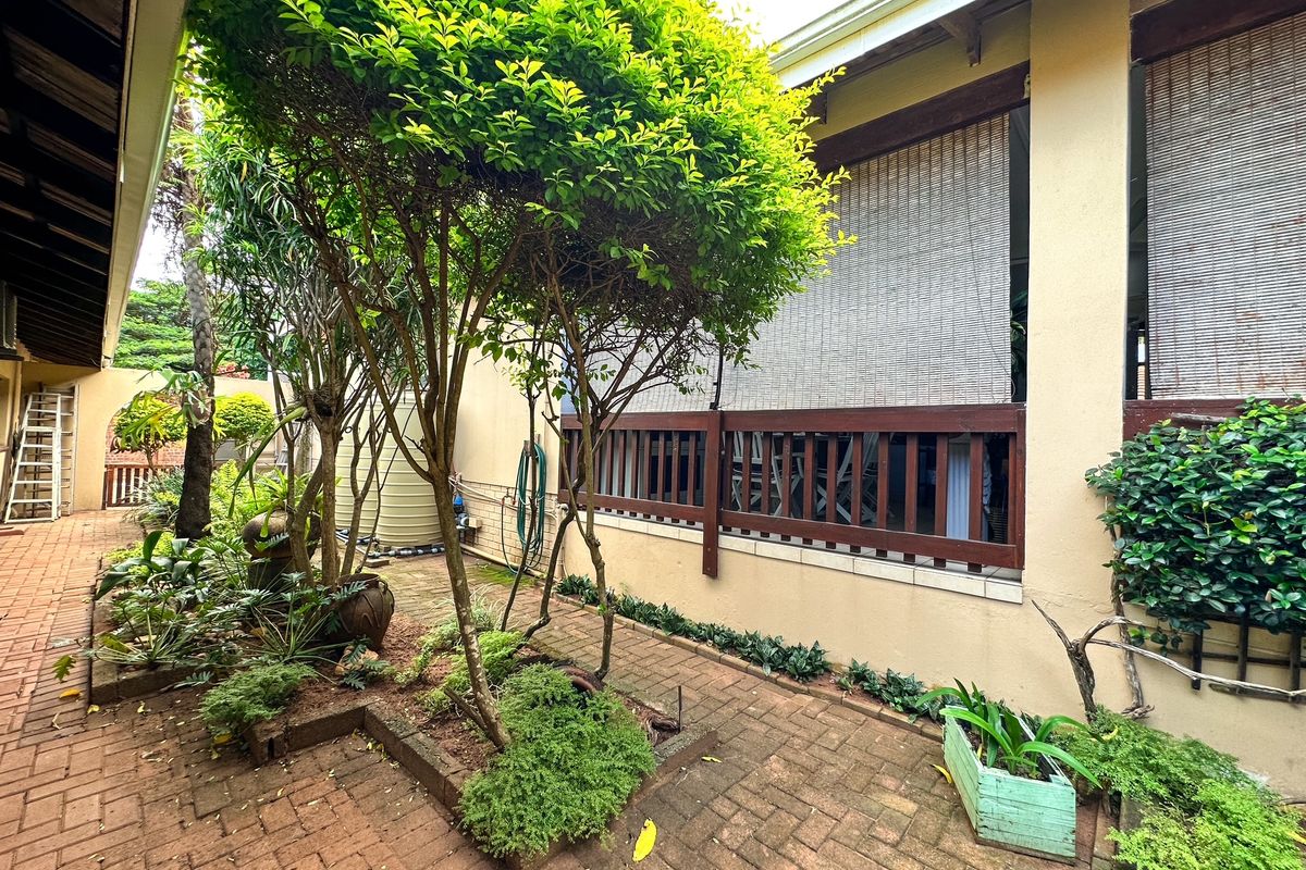 A private side courtyard featuring brick paving, established garden beds, and a covered patio area with timber balustrades and roll-down blinds.